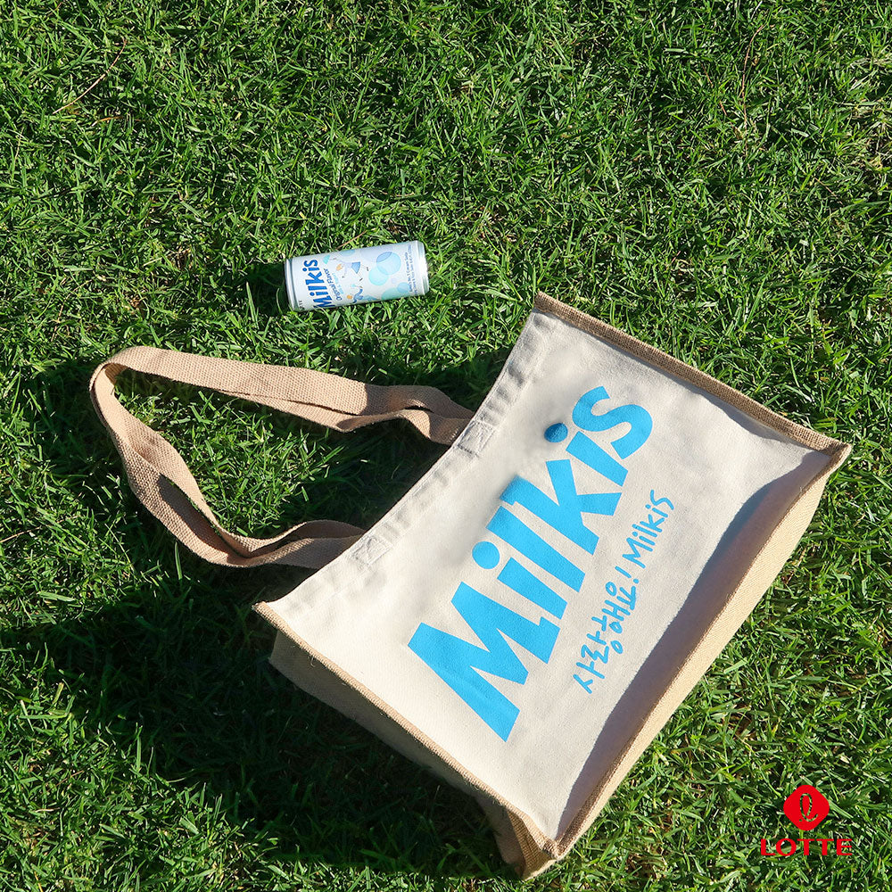 Milkis beige tote bag with blue logo placed on grass, shown with a Milkis original drink can outdoors
