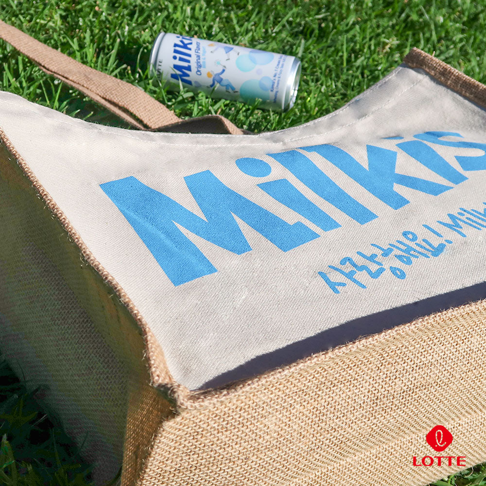 Close-up of Milkis beige tote bag with blue logo outdoors, shown with a Milkis original drink can on grass