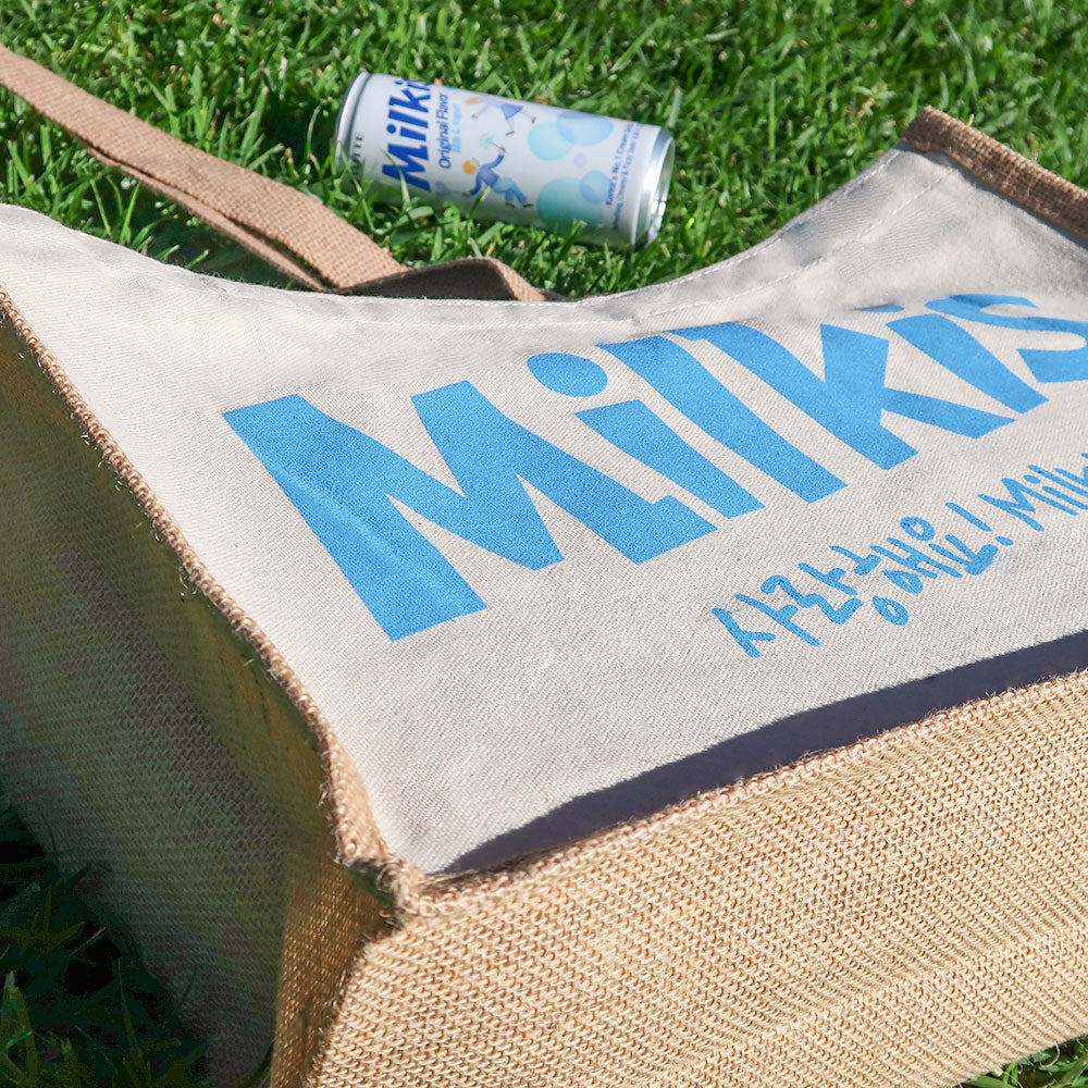 Close-up of Milkis beige tote bag with blue logo outdoors, shown with a Milkis original drink can on grass