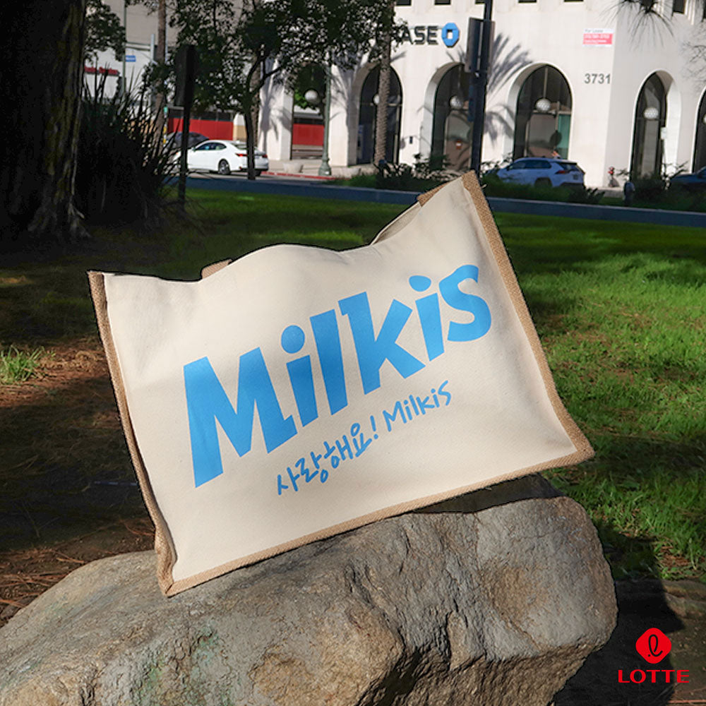 Beige Milkis tote bag with blue logo displayed on a rock in a grassy outdoor setting.